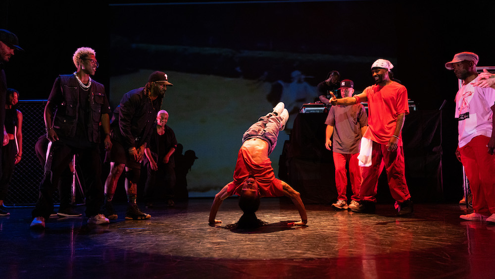a group of dancers in t shirts and sweats in a circle watch as a man with long braids seems to balance upside down on his arms while bending his legs away from his torso. an amazing feet of strength